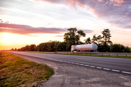 A truck with a semi-trailer tanker transports dangerous goods against the backdrop of a sunny sunset and the sky in the clouds. Transportation of fluid and liquid cargo in cargo transportationの写真素材