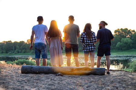 A company of young people stands on the sandy bank of the river and looks at the sunset. Outdoor recreation in summer.の写真素材
