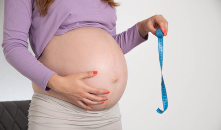 A pregnant girl with a big belly holds a measuring tape on a white background. The concept of measurement and size of the abdomen during pregnancy. The volume of the abdomen by week, intrauterine infectionの写真素材