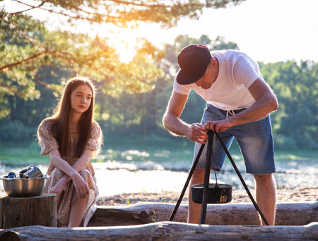 A young guy hangs a bowler hat over a fire against the background of the forest and nature. The guy and the girl are resting on the sandy bank of the river.の写真素材