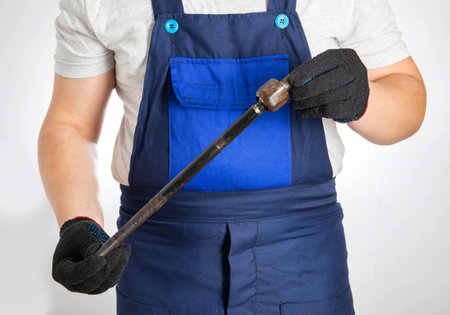 A car mechanic holds a broken car tie rod against the background of a blue overalls. Replacement of steering rods and tips at a car serviceの写真素材