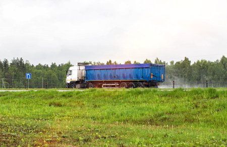 A truck with a semi-trailer for bulk cargo transports sand on a highway in the rain