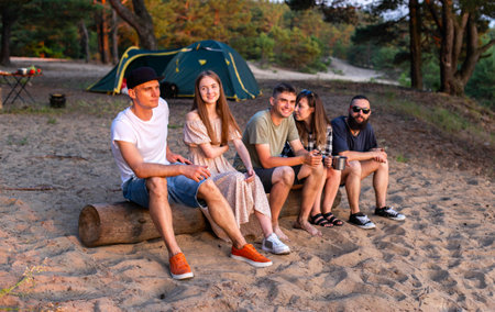 A company of friends guys and girls sits on a log on the river bank in summer. Camping, sandy beach. Adventureの写真素材