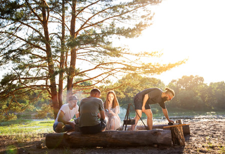 Smiling guys and girls are resting in the summer on the banks of the river with a tent. Cooking food on a fire. Hiking. Beautiful summer nature.の写真素材