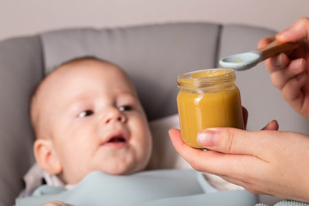 Mother holds a jar of vegetable puree against the background of a 6-month-old baby. First food for babies.の写真素材