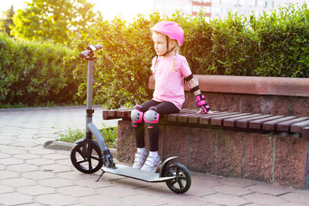 A beautiful seven-year-old girl sits on a bench in pink protective equipment next to a scooter. The concept of cycling and scootering without injury.の写真素材