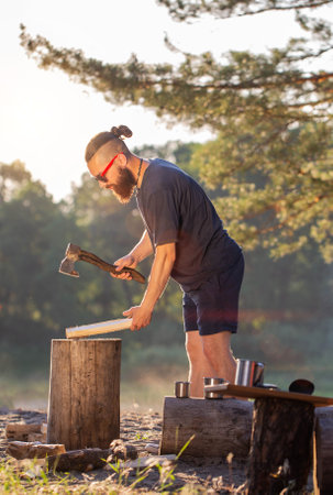 A stylish brutal man with a beard and black glasses is chopping wood on a campsite to kindle a fire....