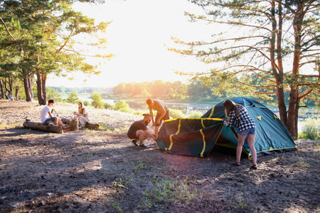 A company of friends guys and girls set up a tent on the sandy bank of the river in summer. Hiking, outdoor recreation in the forest. Sunny day, countrysideの写真素材