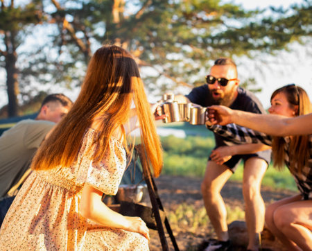 The company of girls and guys celebrate a holiday in nature. They clink glasses with drinks. Rest in...