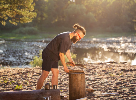 A brutal man with a beard and an ax in his hands is chopping wood for a fire at a campsite. Outdoor recreation in summer on the banks of the river. Bushcrafting.の写真素材