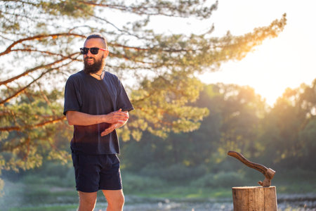 Handsome perfect man with a beard and black glasses outdoors in summer against the backdrop of a forest and a river bank. The concept of outdoor recreation, survival in nature. Bushcrafting.の写真素材