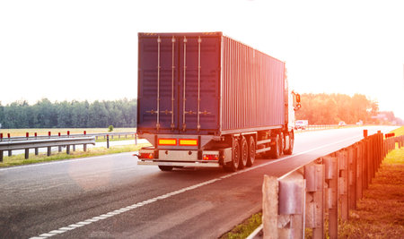 A container truck with a semi-trailer transports cargo from a port to another country along a highway in the summer against the backdrop of the sun. Logistics in cargo transportation, businessの写真素材