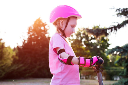A beautiful girl in a pink protective outfit rides a scooter through the streets of the city in summer. Safe cycling and scootering without injury, movementの写真素材