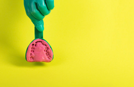 The hand of a dentist doctor in a green medical glove holds a dental spoon with an alginate plastic mass for making a plaster cast of the patient s jaw on a yellow background. Concept of malocclusion, installation of braces, production of dentures. Copy space for text, problemの写真素材