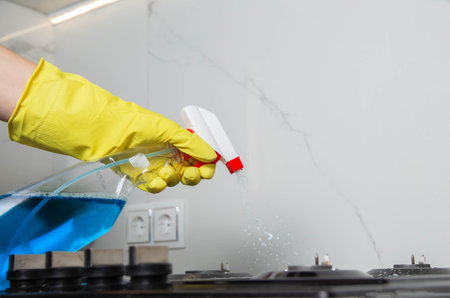 A girl's hand with a cleaning spray sprays on a built-in gas stove. Modern anti-grease, antistatic cleaning product. Background white porcelain tiles on the wall. Copy space for textの写真素材