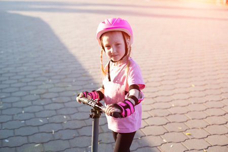 A beautiful girl of 7 years old in a pink helmet and armlets stands on a scooter. Summer sunny day.の写真素材