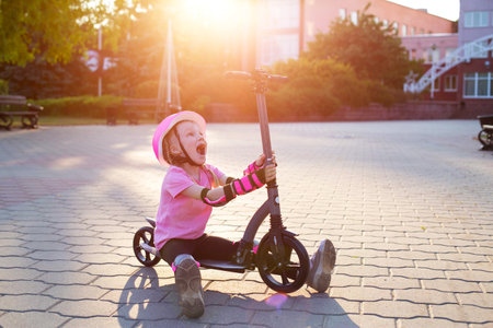 A beautiful caucasian girl of seven years old in a pink protective helmet and outfit is fooling around on a scooter in the summer. Smiling girl laughs next to the scooter.の写真素材
