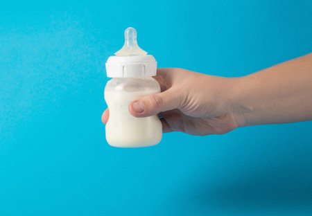 Father's hand with a baby bottle with milk formula on a blue background shakes the mixture to mix....