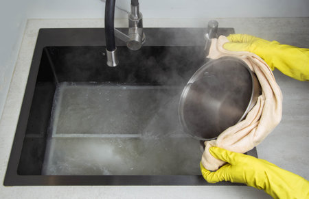 Man's hands in yellow gloves pour pots of hot water into a sink with dirty filters from the hood in the kitchen. Cleaning from dirt and grease with an oxygen cleaner.の写真素材