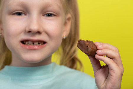 Beautiful little girl eating a chocolate bar on a yellow background. Sweet food, calories. Copy...