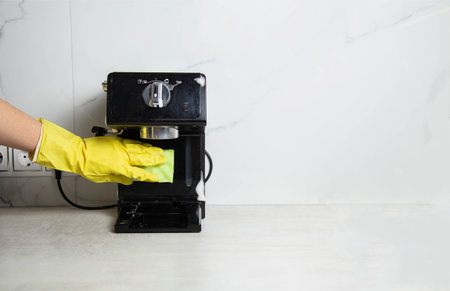 A girl s hand in a yellow glove washes a black coffee machine with a sponge with foam and cleaning agent in the kitchen against a background of a white wall with porcelain tiles. Copy space for textの写真素材