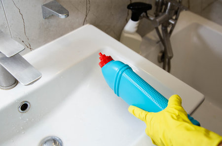 The hands of a person in a yellow cap with a blue bottle of detergent clean and wash the sink from limescale and dirt. Sanitary equipment washing. Copy space for text, residueの写真素材