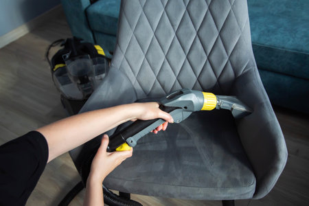 The hands of a girl with a washing vacuum cleaner clean and wash the gray upholstery of a soft chair from dirt. Cleaning services, business. Copy space for textの写真素材