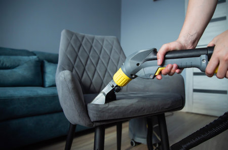 The hands of a girl with a washing vacuum cleaner clean and wash the gray upholstery of a soft chair from dirt. Cleaning services, business. Copy space for textの写真素材