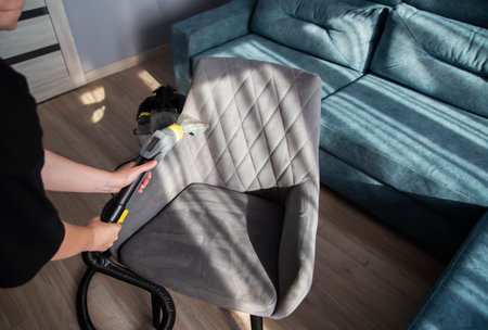 A girl's hand with a washing vacuum cleaner cleans the gray upholstery of a fabric chair in the...