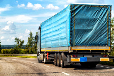 A truck with a semi-trailer drives along a highway against the backdrop of a forest in summer....