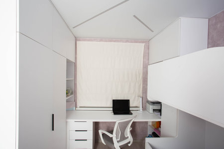 Modern workplace with laptop, computer chair and table in children's bedroom. White furniture and roller blinds on the window, visual comfortの写真素材