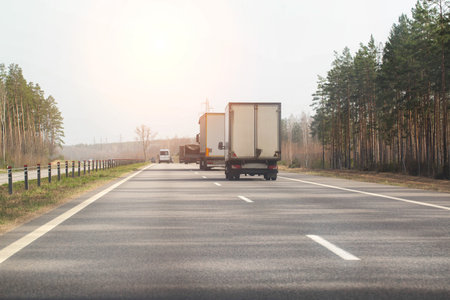 Traffic from trucks transporting goods along a country asphalt road against the backdrop of forest and sun in spring. Economics and business of cargo transportation. Copy space for text, industryの写真素材