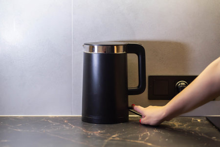 Female hand turns on black modern electric kettle against gray wall in kitchen. Copy space for text,...