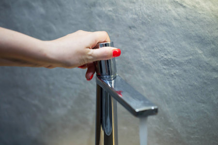 Female hand with red manicure turns on water in modern chrome faucet with floating head
