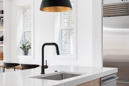 A kitchen sink detail with a black faucet under a black and gold pendant light, white oak island, white countertop, and decorations in the background.の写真素材