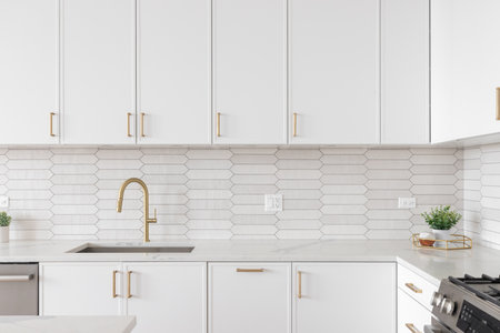 A beautiful kitchen faucet detail with white cabinets, a gold faucet, white marble countertops, and a brown picket ceramic tile backsplash.の写真素材