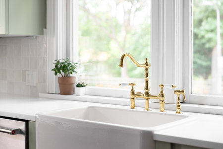 A kitchen faucet in a cozy green kitchen with a gold bridge faucet, white apron sink, tiled backsplash, and white marble countertop.の写真素材