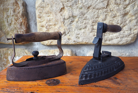 two old rusty antique iron with wooden handle on old wooden desk with sandstone wall backgroundの写真素材