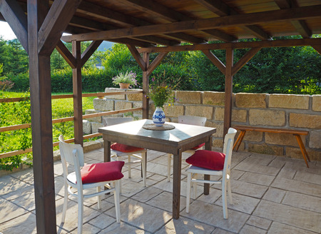 brown tiber wooden gazebo - pergola with table and chairs with red pillows with sandstone wall and floor pavement with summer flowers in the gardenの写真素材