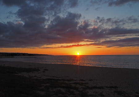 sunset with the red orange sun going down to the sea on the sand beach with dark cloudesの写真素材