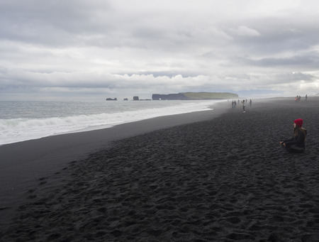 South Iceland, Vik i Myrdal, July 4, 2018: Young pretty woman in red hat sitting meditating or relaxing on Reynisfjara black sand beach, Dyrholaey rock and group of tourist people in backgrund, fog and moody sky, monochromatic lookのeditorial素材