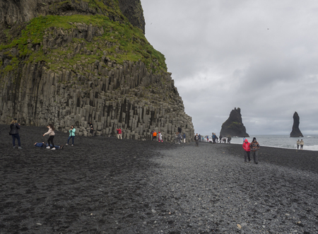 South Iceland, Vik i Myrdal, July 4, 2018: Group of colorful dressed tourist people takeing pictures and havenig fun at Basalt rock columns and Troll toes on black sand beach reynisfjara, fog and moody sky, monochromatic lookのeditorial素材