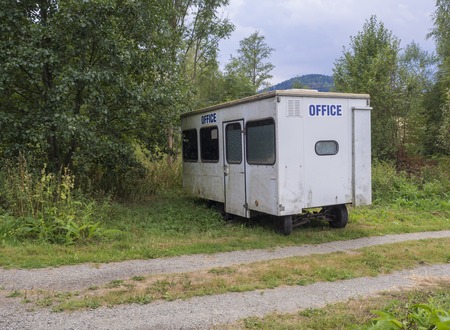 Small white trailer house with blue sign office, abandoned caravan in rural landscape near footpath,...