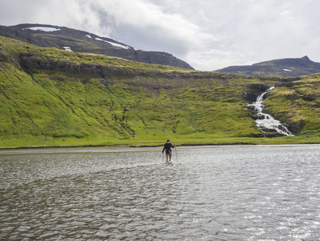 Hiker woman with trekking poles in jacket, purple hat and bare legs crossing river in Iceland Hornstrandir on trek to Hornbjarg cliff, waterfall and green hills in background. Hiking and leisure themeの写真素材