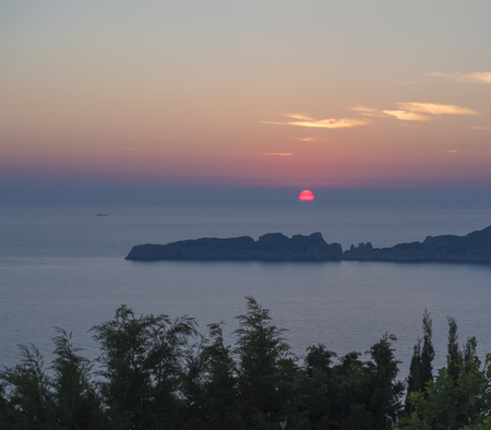 Beautiful colorful orange sunset sun falling to the ocean with rocks in mediterranean sea on Porto Timony, green bush in foreground Corfu,island, Greece.の写真素材