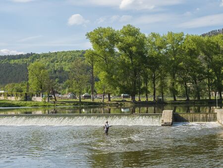 Weir on the river Berounka in village Zadni Treban with fly fisherman standing in the stream and lush green trees. Sunny summer day, Czech Rupublicの写真素材