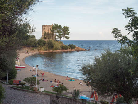 Santa Maria Navarrese, Sardinia, Italy, September 6, 2020: view of Spiaggia di Santa Maria Navarrese beach with group of relaxing people, green trees and old stone tower. Golden hour lightのeditorial素材