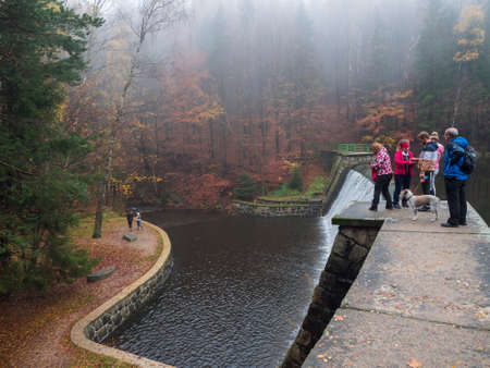 Czech Republic, Nadeje, November 1, 2020: Group of people, family on walk with dog standing on a dam at forest lake with autumn colorful trees at Luzicke hory Lusatian Mountainsのeditorial素材