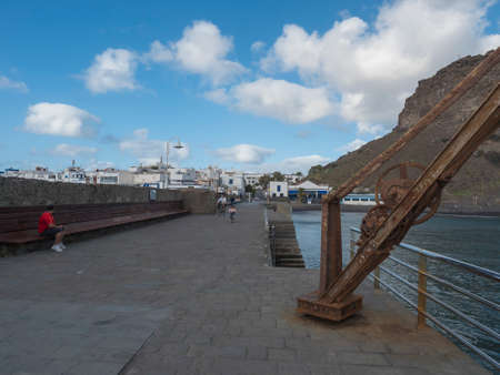Puerto de las Nieves, Agaete, Gran Canaria, Canary Islands, Spain December 17, 2020: Promenade street by sea with rusty crane and Roque de las Nieves rock at traditional fishing villageのeditorial素材