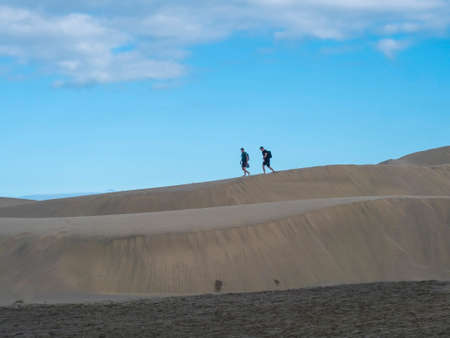 Maspalomas, Gran Canaria, Canary Islands, Spain December 18, 2020: Two barefoot man couple walking at the edge of sand dune at Natural Reserve of Dunes of Maspalomas, blue sky backgroundのeditorial素材